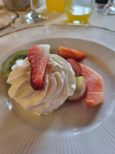 The image displays a close-up shot of a dessert or breakfast item arranged on a white, possibly ceramic, plate. The central focus is a generous swirl of fluffy, white whipped cream. Surrounding and partially nestled within the whipped cream are various pieces of fresh fruit.

To the left, a prominent slice of bright red strawberry with visible seeds is positioned near the base of the whipped cream. Another strawberry slice is visible further up and to the left. Below the whipped cream, a slice of green kiwi fruit is peeking out. Towards the top of the arrangement, there are several slices of other fruits: a piece of pink-orange fruit, possibly guava or papaya, is visible at the very top, and below it, a slice of red-skinned fruit, possibly an apple or another berry, is present.

The plate itself is white with a distinctive ridged or fluted pattern around its rim, creating a textured surface that catches the light. The lighting suggests an indoor setting, possibly with artificial light, as there are reflections on the plate and in the blurry background.

The background is out of focus, indicating a shallow depth of field, which draws attention to the food. In the blurred background to the left, a glass containing a bright yellow liquid, likely orange juice, is visible, along with hints of other tableware and sparkling reflections, suggesting a dining environment such as a restaurant, hotel, or home.

No text is visible in the image. The overall impression is of a fresh, appealing, and well-presented dish, ready for consumption.