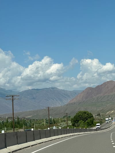 The image depicts a highway scene in Kok Moynok Eki, Kyrgyzstan, under a bright blue sky with scattered white clouds. The road curves to the right, with a concrete barrier separating it from the surrounding landscape. To the left of the road, there are utility poles with wires, a field of green vegetation, and distant mountains with dry, barren slopes. On the right side of the road, there are more utility poles, a dense green tree, and several white vehicles, including trucks and vans, driving on the highway. The overall impression is of a rural highway environment with mountainous scenery.