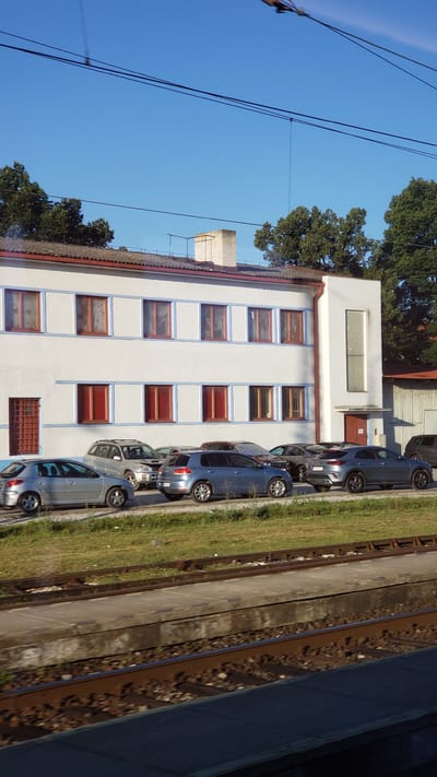 A long shot shows a white industrial building with blue trim and numerous windows with red frames. A parking lot in front of the building is filled with several cars. The building is set back from a train track and a grassy area. Trees are visible behind the building and to the right. The sky is clear and blue.