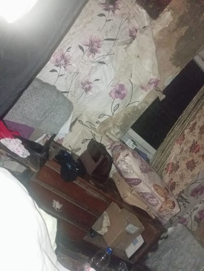The image depicts a cluttered interior space, possibly a bedroom, with items strewn about. A large piece of furniture, possibly a dresser, is visible with drawers. Several fabric items are present, including a gray knitted item, a pink and white patterned fabric, and curtains with floral and butterfly motifs. A black shoe with "AC" written on it is on the floor. A cardboard box with labels is also visible. The lighting appears dim, suggesting it is nighttime or the room is poorly lit. There is no indication of people or specific actions taking place. The location is identified as Rahachow, Belarus.