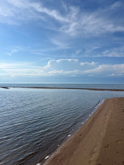 A wide-angle, eye-level, vertical shot captures a serene beach scene under a bright blue sky filled with wispy clouds. The foreground is dominated by a sandy beach sloping gently towards the water. The water itself is calm, with gentle ripples reflecting the sky. A narrow strip of land, appearing to be a sandbar or a causeway, extends across the water in the mid-ground, creating a distinct division. In the distance, the horizon is a soft line where the water meets the sky, with faint mountains visible on the far left. The overall impression is one of tranquility and natural beauty, consistent with a location like Ak Dobo, Kyrgyzstan, known for its natural landscapes.