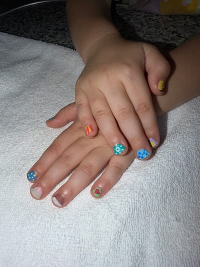 A close-up, overhead shot shows a child's hands with brightly painted fingernails resting on a white, textured towel. The nails are decorated with various patterns and colors, including yellow with black dots, blue with yellow dots, green with white dots, pink and yellow stripes, and a nail with a red heart on a green background. The lighting suggests an indoor setting. No specific location in the city of Urusovo, Russia, can be identified from the image.