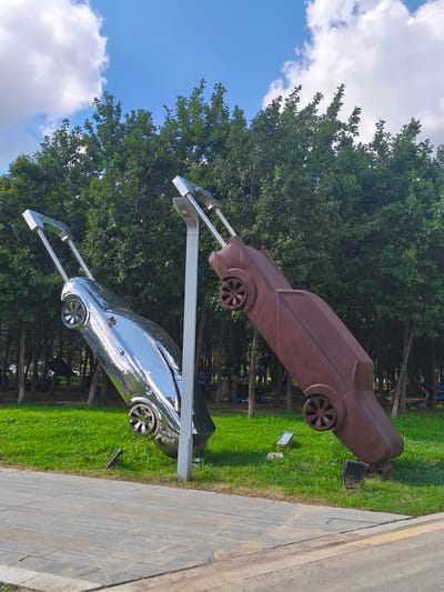 Two car sculptures are displayed in a park setting, against a backdrop of lush green trees under a bright blue sky with white clouds. The sculpture on the left is made of polished chrome, reflecting the surroundings. The sculpture on the right appears to be made of rusted metal. Both sculptures are positioned as if in motion, tilted upwards, with their wheels visible. They are situated on a grassy area next to a paved pathway. In the background, several people can be seen relaxing or walking amongst the trees. This scene is located in Nanguan District, China.