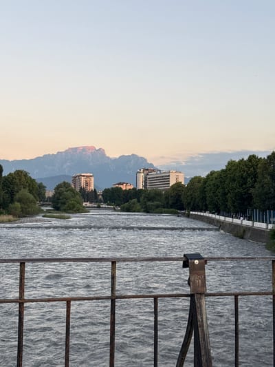 The image depicts a waterfront scene with a river in the foreground, its surface textured with ripples and small waves. A rusty metal railing with vertical and horizontal bars runs along the right side of the frame, partially obscuring the view of the river. On the left side of the river, a paved walkway lines the bank, bordered by lush green trees. Behind the trees, several multi-story buildings are visible, suggesting an urban or semi-urban environment. In the distance, majestic mountains rise, their peaks bathed in the warm, soft light of what appears to be either sunrise or sunset. The sky transitions from a pale yellow near the horizon to a soft blue overhead, with faint clouds scattered across it. The overall atmosphere is tranquil and scenic, with a blend of natural beauty and man-made structures. The time of day is likely either dawn or dusk due to the lighting on the mountains and sky. No people are clearly visible, and no text is present in the image. The location context provided is Askhazan, Russia, which aligns with the mountainous backdrop.