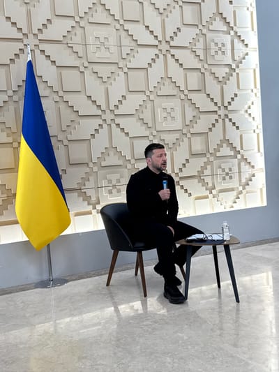 The image shows a man sitting in a black leather chair, speaking into a microphone. The chair has wooden legs. He is wearing a black suit jacket and black pants. He has short brown hair and a beard. He is looking to the right, toward a white wall behind him. There is a small table in front of the chair, to the right, which has a water bottle, a notepad and a pen on it. The man is in front of a wall covered with a pattern of white squares. To the left of him, there is a flagpole holding the Ukrainian flag. The scene appears to be in a government building in Ankara, Türkiye (latitude: 39.851, longitude: 32.827).  The location is most likely the Turkish Grand National Assembly (TBMM).