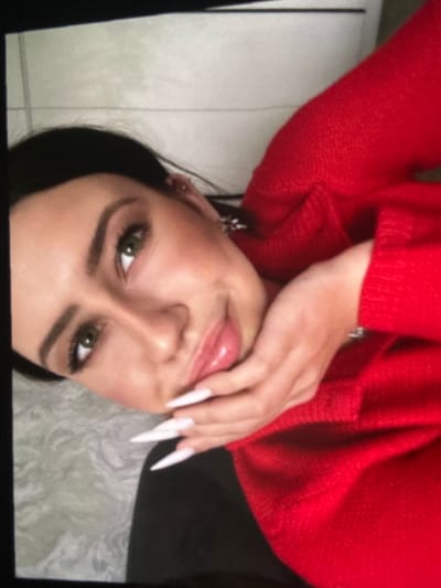 A young woman with dark hair, green eyes, and long white nails is shown from the chest up, looking towards the viewer with her left hand touching her chin. She is wearing a red sweater. The background consists of a tiled surface and a grey patterned textile. The image appears to be a close-up photograph taken indoors. There is no identifiable information within the image to indicate a specific location in Korostyshiv, Ukraine.
