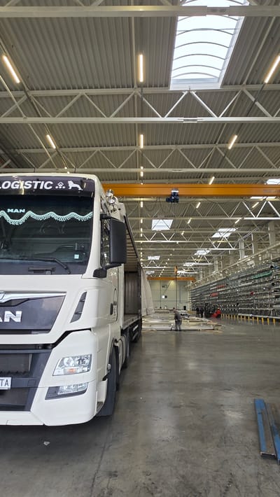 A white MAN truck is parked inside a large industrial building in Gara Elin Pelin, Bulgaria. The building features a high ceiling with exposed steel beams and artificial lighting. An overhead crane runs along the length of the building. A person is standing near the back of the truck, possibly overseeing the loading or unloading process. The concrete floor is a uniform gray. Some metal components or materials are visible in the background.