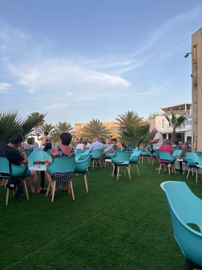 This image depicts an outdoor seating area with people dining and socializing. The area is covered with artificial grass and furnished with aqua-colored chairs around small, circular tables. In the background, buildings with light-colored facades and palm trees are visible under a partly cloudy blue sky. The scene appears to be a restaurant or cafe, likely in Taguermess, Tunisia, given the architecture and vegetation often found in coastal areas of Tunisia. Several individuals are seated at tables, some engaged in conversation, while others are holding drinks. One person stands near a table, holding a phone. The overall atmosphere suggests a relaxed, late afternoon or early evening gathering.