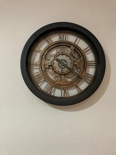 The image shows a circular, steampunk-style wall clock mounted on a white wall. The clock has a black outer rim. The clock face features Roman numerals and exposed, decorative gears. The clock hands indicate a time of approximately 10:12. There are no people, subjects, or interactions visible in the image. The location is not specified, and there is no indication that it is located in Tres de Mayo, Mexico. The environment consists solely of a white wall.