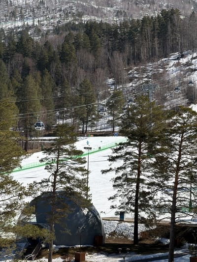 The image shows a ski resort in Озёрное, Russia. A gondola lift carries people up a forested mountain. Skiers and snowboarders are visible on a snow-covered run marked with green fencing. A geodesic dome structure is present near the bottom of the ski run. Trees are in the foreground of the image.