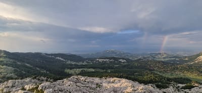 The image is a panoramic landscape view from a rocky, elevated vantage point overlooking a mountainous region. The foreground is dominated by rough, light-colored rock formations, indicative of a karstic landscape. Beyond the foreground, rolling hills covered in dense, dark green forest stretch into the distance. In the mid-ground, there are more rounded mountains, some with visible rocky outcrops.

The sky is dramatic, filled with heavy, grey clouds that appear to be breaking apart in places, allowing shafts of light to pass through. A faint rainbow is visible on the right side of the image, arcing across the sky and disappearing behind a distant mountain range. The overall lighting suggests it is either early morning or late afternoon, with a mix of shadow and illumination across the landscape.

The location is identified as Ubla, Bosnia and Herzegovina, a mountainous area known for its natural beauty. The scene captures a moment of transition, with the moody sky and the presence of a rainbow hinting at recent or impending rain. There are no people or man-made structures immediately visible in the frame, emphasizing the natural and wild character of the environment. The image conveys a sense of vastness and rugged terrain.
