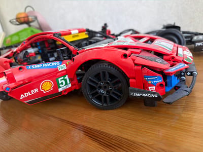 This image shows a red Lego Technic Ferrari race car model resting on a wooden surface. The car is detailed with various stickers representing sponsors and racing numbers, including "SMP Racing", "Adler", "Shell", and the number "51". The car also features a Michelin "V-Power" sticker. The focus is on the front portion of the car, including the wheel, fender, and front spoiler. In the blurred background, another Lego model of a black race car can be partially seen. An apple sits in a wire basket in the background. The location cannot be determined from this image.
