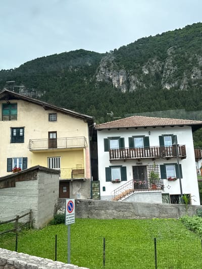 The image depicts two houses situated on a hillside in Fai della Paganella, Italy, with a densely forested mountain slope dominating the background under an overcast sky. The house on the left is painted a pale yellow and features multiple windows with dark green shutters, some of which are open, revealing the interior. A balcony with a wrought-iron railing is visible on the second story, above a lower yellow-painted section. To the left of this house, a smaller, grey concrete structure with a wooden roof extends towards the foreground.

The house on the right is white with dark green shutters on its windows, similar to the neighboring building. It has a prominent wooden balcony with ornate white railings on the upper floor. A set of stairs with a decorative metal railing leads to the entrance on the ground floor, which has a black-grated door. The roof of this house is covered with terracotta tiles.

In the foreground, a lush green lawn stretches out, bordered by a low stone wall and a wooden fence. A signpost with a circular red and blue symbol and Italian text, indicating "private property" and "authorized parking," stands on the lawn. The overall scene suggests a residential area nestled within a mountainous landscape.