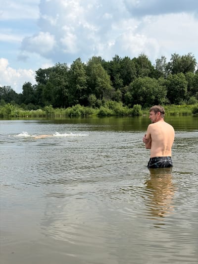 A young adult male, with light brown hair and a muscular build, stands in waist-deep water on a body of water, facing away from the camera and to the left. He is wearing black swim trunks with a palm tree pattern. His arms are crossed in front of his chest. In the distance, to the left of the frame, another person is swimming, creating a splash. The background consists of a lush green tree line that stretches across the horizon, under a partly cloudy sky. The water is calm, with ripples reflecting the sky and surrounding environment. The scene appears to be a natural outdoor setting, likely a river or lake, in the outskirts of Кватчи, Russia, characterized by its abundant vegetation and tranquil water.
