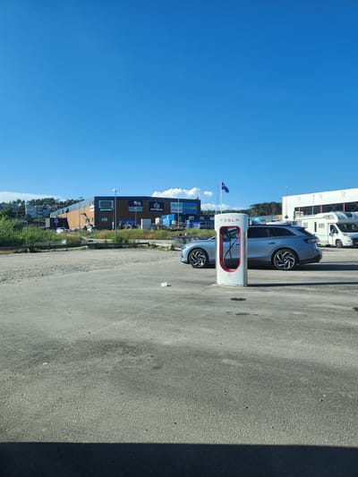 This is a media file showing a Tesla charging station in Strömstad, Sweden. The scene is an outdoor parking lot on a sunny day with a clear blue sky. A white Tesla charging station with red accents is prominently featured in the foreground. A grey Tesla vehicle is parked next to it, plugged into the charger.
In the background, there are several buildings with visible signage. One building has signs for "GÖTTEN," "spg," and "ÖSTERHOLM GARDEN" and "apoteket". Another building has signs for "Eurocash" and "Vassmo". A white RV or campervan is also visible parked in front of one of the buildings. Trees and greenery are present behind the buildings, suggesting a location near natural surroundings. The ground is paved asphalt. The overall impression is a modern, functional setting for electric vehicle charging. There are no people visible in the image. The time of day appears to be midday, given the bright sunlight and distinct shadows.
