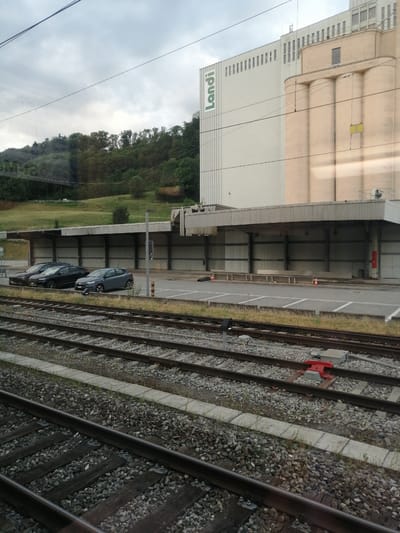 This image captures a view of a commercial building with a large silo structure, likely a warehouse or factory, situated next to a train track. The building features the name "Landi" in large green lettering on its side. Several parked cars are visible in a designated parking area in front of the building, under a covered structure. The train tracks dominate the foreground, with ballast and wooden sleepers clearly visible. The scene appears to be set in an industrial or commercial area, possibly on the outskirts of a town, given the presence of the train infrastructure and the large building. The sky is overcast, suggesting a cloudy or potentially rainy day. The time of day is not explicitly clear but the lighting suggests it is daytime. The image provides a snapshot of a functional, working environment.