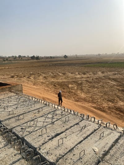 A man in a suit and hard hat walks on a newly constructed concrete structure beside a dirt road. The concrete has a repeating pattern of square shapes with vertical metal bars. A vast, flat field stretches to the horizon under a hazy sky. The image is rotated counter-clockwise.