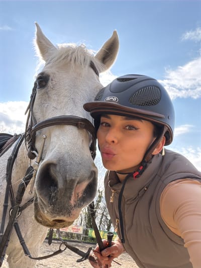A person wearing a riding helmet and a vest is posing with a white horse in what appears to be an outdoor setting. The horse is wearing a bridle. In the background, a sign can be seen with some text, including the word "MEGA". The city and country the image was taken in are Grad Labin, Croatia. The person is holding the reins of the horse.