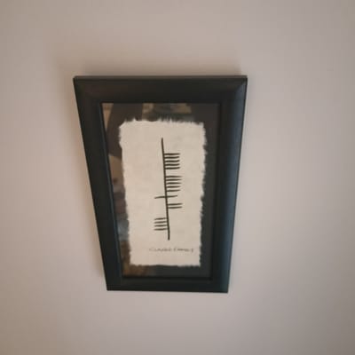 A framed artwork is displayed on a pale wall. The artwork itself is a print on textured, off-white paper, featuring a vertical line with several horizontal strokes to its left and right, arranged in clusters. Below this symbol, there is some faint text that appears to read "CIANN FAMILY". The frame is dark, possibly black or dark wood, with a slightly beveled edge. The image is taken from a slightly upward-angled perspective. The surrounding environment is a plain, neutral-colored wall, with soft, ambient lighting. There are no people visible in the image. The specific location in the city of Limerick, Ireland, cannot be determined from the visual information provided in this image.