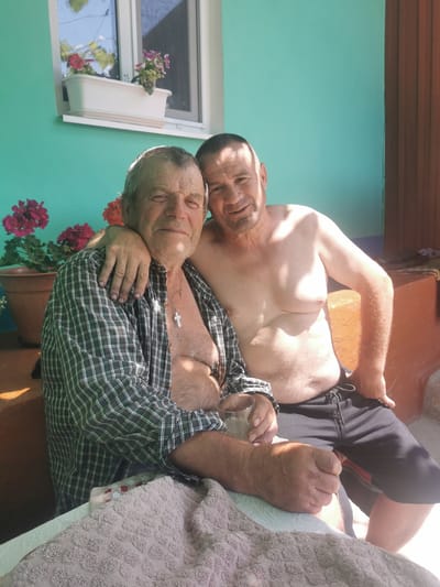 Two men are sitting on a porch outside a house in the village of Cărpuna, Moldova. The man on the left is older, with gray hair and a plaid shirt unbuttoned to reveal his chest. He has a cross necklace on. The man on the right is younger, shirtless, with short brown hair and is wearing black shorts. He has his arm around the older man. Behind them, the house is painted a bright teal color and there is a window with a flower box on the sill. In front of them, a patterned towel is draped over a surface. The lighting suggests it is daytime and sunny.