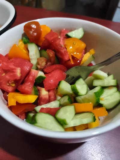 A close-up, eye-level shot shows a white bowl filled with a chopped tomato and cucumber salad. The salad contains red tomatoes, orange bell peppers, and green cucumbers, all cut into bite-sized pieces. A fork is partially visible in the right side of the bowl, with some salad ingredients on its tines. The bowl is set on a dark red surface, possibly a tablecloth or countertop. The background is blurred, but suggests an indoor setting. The scene is devoid of any recognizable landmarks or specific details that would indicate its location in Sokolivske, Ukraine.