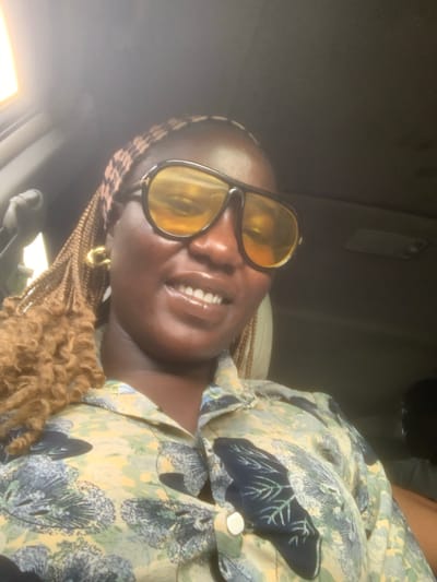 The image depicts a female individual of African descent captured in a chest-up portrait inside a vehicle in the city of Oyigbo, Nigeria. She is smiling, revealing her teeth, and her lips appear glossy. Her hair is styled in light brown/caramel-colored braids with a textured, curly appearance at the ends, and a patterned head covering is visible on her scalp. She wears large, dark-framed aviator-style sunglasses with yellow-tinted lenses, obscuring her eyes. A gold-colored hoop earring is visible on her left ear. She is dressed in a collared button-up shirt featuring a light green/yellow base fabric with a darker green/blue abstract botanical or leaf pattern. The background is predominantly dark and out of focus, indicative of a low-light interior environment. A bright light source, possibly an interior vehicle light, is visible in the upper left corner. The partial head and shoulder of another individual are dimly discernible in the lower right background, suggesting the presence of another passenger. The primary subject is looking directly at the camera.