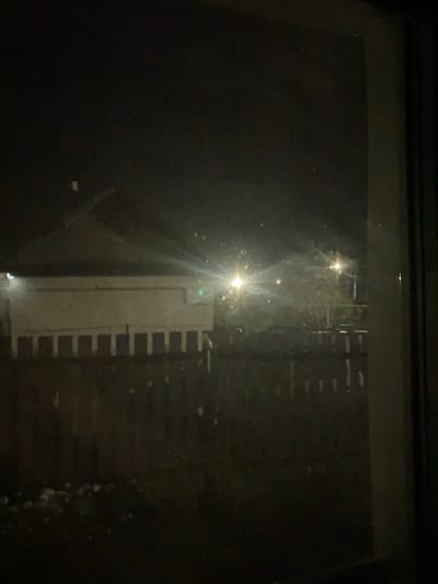 The image is a dark, grainy photograph taken at night through a window, likely from inside a residence. The view outside shows a dimly lit outdoor scene. In the middle ground, there is a light-colored building with a prominent dark, sloped roofline. The building appears to have a row of evenly spaced, dark rectangular openings along its lower facade, suggesting a structure with an arcade or enclosed walkway. In front of this building, there is a dark wooden fence, also with regularly spaced vertical elements. Beyond the fence, trees and vegetation are visible, illuminated by two bright, diffused light sources that appear to be streetlights or security lights. The lights create a lens flare effect and scatter light, contributing to the overall haziness of the image. There are small, bright specks scattered throughout the scene, possibly dust or water droplets on the windowpane or in the air. The foreground is very dark, with some indistinct white or light-colored debris on the ground, and a faint pink object that may be a flag or marker. The overall impression is of a suburban or semi-rural environment at night, with artificial lighting casting a glow on the surroundings. The mention of Shiryaevo, Russia, provides a geographical context for the scene, suggesting a location in that specific region.