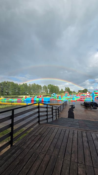 The image captures an outdoor water park located in Jaunmārupe, Latvia. A prominent double rainbow arches across a cloudy sky, with the main rainbow framing a vibrant collection of inflatable water attractions floating on a body of water. These colorful structures, including slides, climbing walls, and various shapes, are scattered across the water's surface. The park appears to be set against a backdrop of trees and a distant shoreline. In the foreground, a wooden deck with a dark railing leads towards the water, providing a vantage point of the park. There are a few individuals visible, some on the inflatable attractions and others near the edge of the water, suggesting the park is operational. The overall scene depicts a recreational area on a potentially overcast day with a striking natural phenomenon.