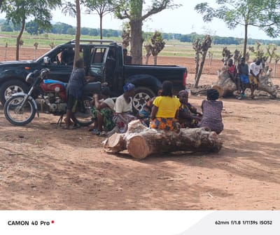 The image depicts a rural scene near Wase, Nigeria, featuring a group of people gathered near a black pickup truck and a motorcycle. Several women are seated on a large, felled tree trunk, engaged in what appears to be conversation or rest. Three young males are sitting on a separate portion of a tree trunk. A young male is leaning on the motorcycle. The environment consists of dirt ground with sparse vegetation, and the landscape shows trees and open fields, indicating a less urbanized area. The overall setting suggests a communal gathering in a rural environment.