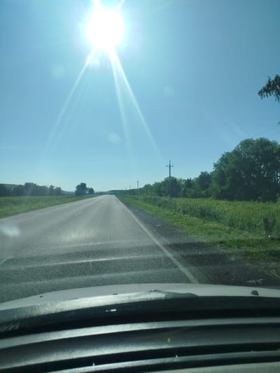 The image shows a view from inside a vehicle driving on a paved road under a bright, clear blue sky. The sun is high and appears to be slightly to the left of the frame, causing lens flare and bright streaks of light. The road is asphalt with a white line marking the right edge. On either side of the road, there are grassy fields and trees. Power lines and utility poles are visible along the right side of the road. In the distance, the road stretches towards the horizon, with a slight curve. The foreground shows the interior of the vehicle, including the dashboard and windshield. The overall impression is of a sunny day drive through a rural or semi-rural landscape. No specific details about the city of Tushna, Russia are visible in the image.