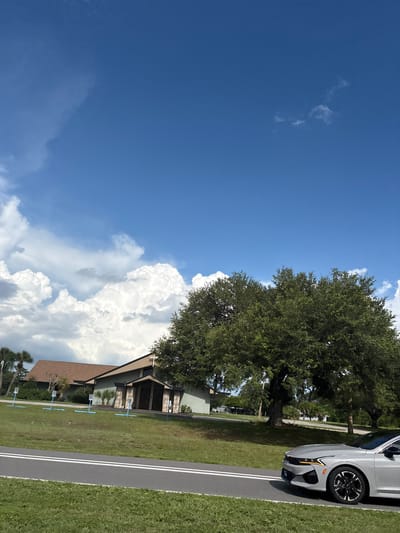 An exterior, ground-level, eye-level shot captures a scene in North Port, United States of America, on a bright, cloudy day. In the foreground, a modern silver car is parked on an asphalt road with white lane markings. Behind the road, a well-maintained grassy lawn slopes up towards a light-colored, single-story building with a dark-pitched roof. The building features a prominent entrance with two doors, flanked by stone pillars and a cross at the top. Several blue signs with white markings are visible on the lawn in front of the building, spaced evenly apart. To the right of the building, a large, mature tree with dense green foliage dominates the middle ground. The background is filled with a bright blue sky punctuated by large, fluffy white clouds, some of which are illuminated by the sun. Palm trees and other vegetation are visible on the left side of the building.