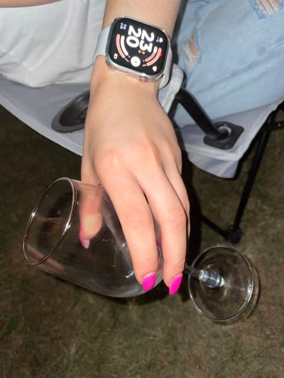 The image shows a close-up of a person's hand and arm, wearing a smartwatch and holding a wine glass. The hand is positioned to pour liquid from the glass. The person is wearing light blue ripped jeans and a white top. The background appears to be outdoors with a grassy or earthy surface, and the dark shadows suggest it might be evening or night. The smartwatch displays the time as 8:53 PM and the date as Tuesday, June 20th. This scene is not specifically located in Jablonné nad Orlicí, Czechia, as there are no identifying landmarks or context provided in the image to suggest this location.