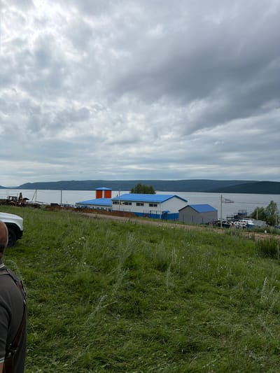 The image shows a cloudy day with a vast body of water in the background, likely the Volga River, as suggested by the context of Sergeevka, Russia. The shoreline is lined with a few industrial buildings featuring blue roofs and some orange accents. In the foreground, a grassy field with scattered wildflowers slopes down towards the water. A person is partially visible on the left edge of the frame, facing away from the camera. Several vehicles, including a white SUV and construction equipment like a backhoe, are present near the buildings. A boat is also seen on the water. The overall scene suggests a riverside industrial or port area with ongoing construction or development.