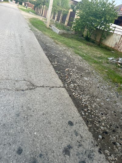The image depicts a street scene in Apsheronsk, Russia. In the foreground, a paved road with visible cracks runs diagonally across the frame. Adjacent to the road is a gravelly shoulder with scattered rocks and debris. Beyond the shoulder, a patch of dry grass is visible. In the background, a beige concrete fence runs along the property line, bordered by a rusty metal fence. A dense green tree with full foliage stands to the right of the fence, casting shadows on the ground. Further in the distance, the rooflines of houses and another fence with brick pillars are visible. Several people, appearing as small figures, are walking on the road in the far distance.