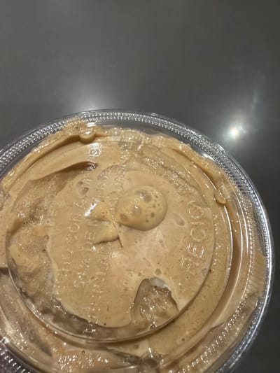 A close-up image depicts the top of a clear plastic cup containing a light brown, creamy beverage. A clear plastic dome lid covers the cup, with the beverage having partially overflowed onto the lid's exterior surface. Bubbles are visible within the spilled liquid, notably a cluster near the lid's center-right. Embossed text is present on the lid, partially obscured by the beverage, with "RECYCLE" discernible on the right and "MADE WITH" visible on the left. The background is a dark, smooth surface, likely a countertop, exhibiting reflections of ambient light. The scene is located in Fraccionamiento Los Heroes Queretaro, Mexico.