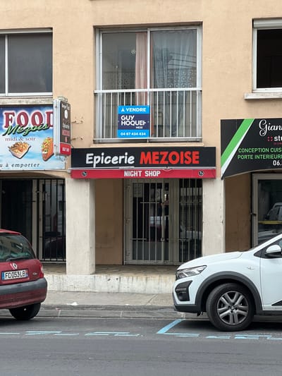 The image shows the storefront of a convenience store called "Epicerie Mezoise," located at 43.428 latitude and 3.606 longitude in Mèze, France.  The storefront is bordered by a red awning and the sign, which also advertises that it is a "Night Shop". The storefront has a glass door and a window that are each covered by a metal security gate. Directly above the door and window is another sign which states "A VENDRE GUY HOQUEH MEZE 04 67 434 434". The storefront is surrounded by a sidewalk and street.  A white car is parked in front of the storefront. To the left of the storefront, a red car is also parked, partially visible.  On the right of the storefront is a sign with faded black letters on a green and white background reading: "Gianni Studio La creativite en obra Conception Cuisine Porte Interieur 06."  Above the "Epicerie Mezoise" storefront is a window that displays a sign with the name "Food Mezois midi & soir".  There is also a smaller sign advertising "Epicerie." There are no people visible in the image.