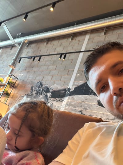 An adult male captures a close-up image of himself and a young child within an indoor establishment in Bretton, United Kingdom. The male, partially visible on the right with dark hair and a light-colored t-shirt, holds the camera, capturing his face from the eyes down, looking directly forward. To his left, a young child with light brown hair styled in multiple braids, some obscuring her face, appears to be sleeping or resting. She is wearing a pink, possibly knitted, garment. Both individuals are seated on a brown, upholstered booth or bench.
The background features an industrial-style interior. The ceiling is gray with exposed silver-colored metallic ductwork running horizontally and vertically. Multiple black track lights illuminate the space. The wall behind them is composed of light gray rectangular blocks, possibly concrete, and features a large, monochrome mural depicting a historical or maritime scene, including what appears to be a horse and parts of a boat. A black metal shelving unit with greenery is visible in the far left background.