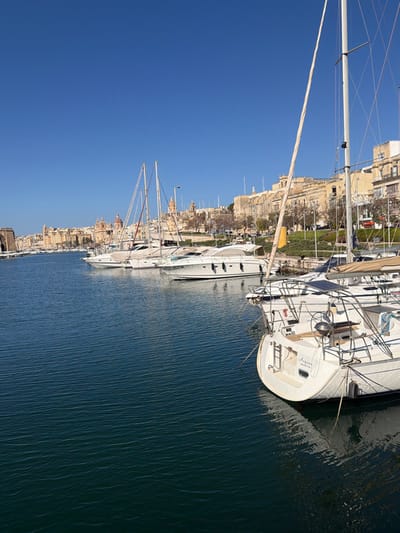 The image displays a marina located in the city of Cospicua, Malta. The foreground features the stern of a white sailing yacht, partially visible on the right, displaying a transom ladder and the inscription "Acquina Malta." Mooring lines are attached to the vessel. The water is dark blue and exhibits slight ripples. Extending into the midground, multiple white recreational vessels are moored, including several sailing yachts with their masts upright and various motor yachts. The background reveals a historic cityscape constructed from light-colored limestone buildings, characterized by numerous domes and steeples, stretching along the waterfront. A fortified structure or extensive building complex is visible on the elevated shoreline. Sparse trees are present along the water's edge in the background. The sky is clear and bright blue, indicating sunny weather. No individuals are visible in the scene.