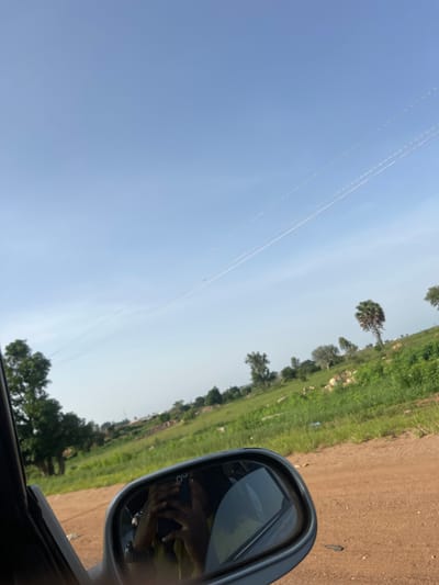 A clear, bright day in Ladura Foron, Nigeria, is captured through the perspective of a car's side mirror, reflecting the driver. The mirror's frame is visible on the left side of the image. The driver, a person of color, is holding a smartphone and taking a selfie, their hands and face partially visible in the reflection. The background shows a vast, green landscape with rolling hills, dotted with scattered trees and some rocks. A dirt road runs through the foreground. In the distance, power lines stretch across the clear blue sky, with a few wispy clouds. The overall scene conveys a sense of travel and observation of the Nigerian countryside.
