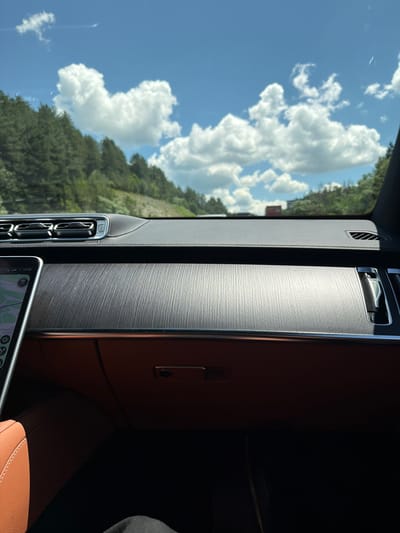 The image is taken from inside a car, looking out the windshield. The dashboard of the car is visible in the foreground, with a textured wood-grain panel. A portion of a tablet device is seen on the left side of the dashboard, displaying a navigation map. Through the windshield, a bright blue sky with fluffy white clouds is visible, along with a dense forest on a hillside. In the distance, a few buildings can be seen. The context of Studena, Bulgaria, is not directly depicted in the image itself, but it is provided as additional information about the location.