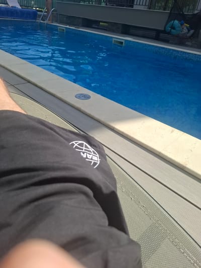 A person is relaxing by a swimming pool. The pool is filled with bright blue water, and there are tiled steps leading into it. The poolside is paved with light-colored tiles and then a section of grey composite decking. A depth marker on the pool edge indicates "1.60 M". The person is wearing black shorts with a white circular logo on them. The image appears to have been taken on a sunny day, with harsh shadows indicating it is likely mid-day. The scene suggests a vacation or leisure setting, possibly a resort or private residence. The overall mood is relaxed and summery.