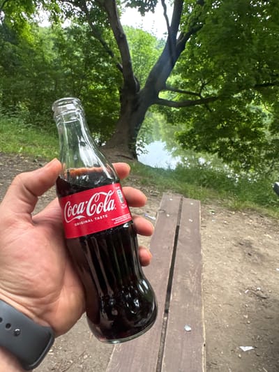 A person's hand is holding a small glass bottle of Coca-Cola. The bottle has a red label with the Coca-Cola logo and the text "ORIGINAL TASTE." The hand is wearing a dark smartwatch on the wrist. The background shows a park-like setting with lush green trees, a body of water, and a wooden bench. The ground is dirt and covered with some small debris. The location is specified as Straszyn, Poland.