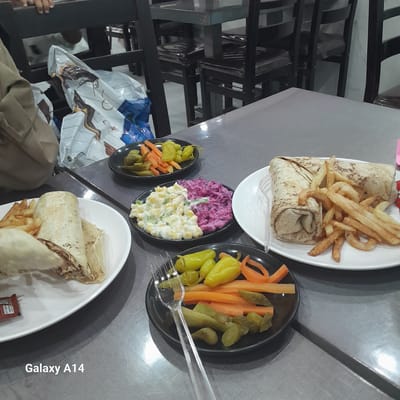 The image shows a table in what appears to be a restaurant or food establishment in Nuseirat, Palestinian Territories. Two plates of shawarma with fries are visible, accompanied by smaller plates containing pickled vegetables (carrots, cucumbers, and peppers) and salads (coleslaw and possibly a macaroni salad). A person is seated at the table, wearing a brown jacket. Plastic cutlery is present. The environment suggests a casual dining setting.