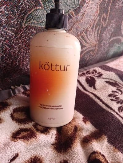 A full shot shows a beige pump bottle with an orange gradient label on a patterned surface. The bottle's label reads "kòttur" in dark orange, with smaller text below in white that appears to be Russian. The text reads "крем с мочевиной с эффектом заботы" and "500 мл". The bottle is positioned on a soft, possibly plush, surface with a leopard-like print pattern. Behind the bottle, the surface transitions to a darker, textured material with a floral or abstract pattern. The lighting suggests an indoor setting, possibly a home environment. The image's composition is centered on the bottle, with the patterned surfaces serving as a backdrop and foreground. No people or actions are depicted.