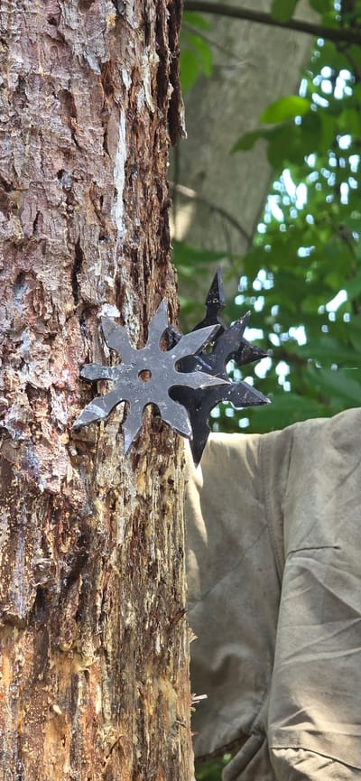This image, observed in the city of Partizanin, Bulgaria, depicts a close-up, vertical view of a tree trunk with multiple throwing stars (shuriken) embedded within its bark. The most prominent throwing star, situated in the mid-left, is six-pointed, dark gray, and features a central circular perforation. Partial text "MADE IN C" is visible on its surface. Behind and to the right of this star, at least two additional, darker-toned throwing stars are partially visible, also lodged in the bark. The tree trunk exhibits thick, fissured, reddish-brown bark, with substantial amounts of white and yellowish resin or sap adhering to its vertical crevices. The background, to the right of the tree, comprises brightly lit, out-of-focus green foliage displaying distinct circular bokeh. Below the foliage, a segment of light tan textile material, possibly a piece of clothing or canvas, with visible seams and folds, is present. No individuals are depicted in the scene.