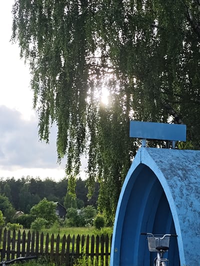 The image depicts a rural or suburban scene, likely in the city of Sudak, Russia, characterized by lush greenery and rustic elements. In the foreground, a bright blue arched structure is visible, possibly a shelter or a decorative element, with a bicycle parked inside. The structure is topped by a rectangular blue sign. Behind the blue structure, a weathered wooden fence runs across the frame. Beyond the fence, a grassy field is dotted with trees and houses, suggesting a residential area interspersed with nature. A dense forest forms the backdrop, and the sky above is partly cloudy with the sun shining through the leaves of a large tree, creating dappled light. The overall impression is one of a tranquil, natural setting on a partly sunny day.