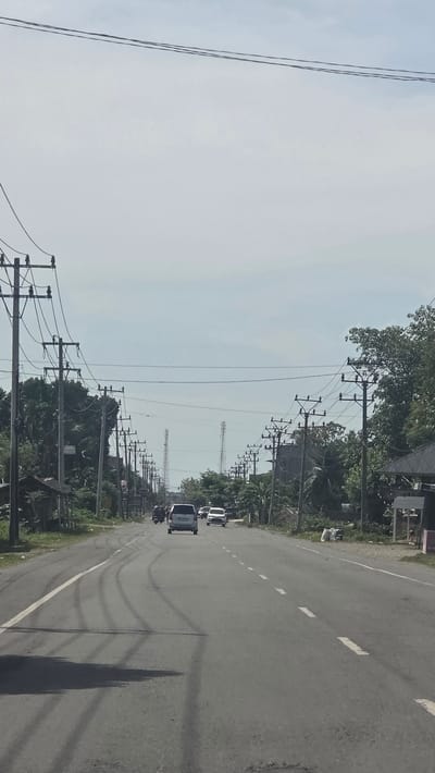 A road scene, likely in Teungoh Baro, Indonesia. The road extends into the distance with a line of utility poles on either side. There are several vehicles on the road, including vans and a motorcycle. The sky is overcast. The road is marked with dashed white lines. Shadows suggest the time of day. Vegetation is visible along the road.