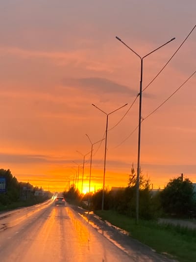The image captures a road in Nurkeevo, Russia, during a sunset. The sky is a gradient of orange and pink, with clouds illuminated by the setting sun. A car is visible driving away from the camera down the wet asphalt road, its taillights reflecting in puddles. Numerous streetlights line the right side of the road, their poles and wires silhouetted against the colorful sky. Trees and vegetation are present on the sides of the road, with some buildings or structures visible in the distance. The wet road surface suggests recent rain or ongoing precipitation.