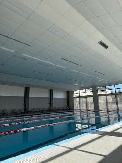 This image depicts the interior of a modern, multi-lane indoor swimming pool facility in Dzhebel, Bulgaria. The large rectangular pool, filled with clear blue water, features at least four lanes demarcated by red and white floating lane lines. The pool deck is tiled with light grey squares, illuminated by natural light entering through a wall of tall, segmented windows on the right. Strong sunlight casts defined shadows onto the deck.
Along the left-hand wall, a row of approximately twelve dark-colored lounge chairs is neatly arranged on a tiled surface adjacent to the pool. The lower portion of this wall is dark grey, while the upper section is a lighter textured grey; dark vertical columns are interspersed along its length. The ceiling consists of a grid of white modular panels, integrating numerous rectangular LED light fixtures, some of which are illuminated, and several dark rectangular ventilation grilles.
Beyond the expansive windows, parts of a multi-story building with numerous windows are visible, suggesting an urban or institutional setting. There are no individuals present in the scene, and no actions or interactions are occurring. The environment appears clean, well-maintained, and unoccupied.