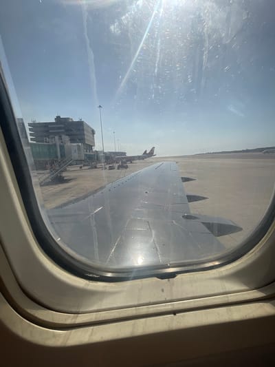 View from an airplane window showing the wing of the plane, the tarmac, and an airport terminal building. The location in Catia La Mar, Venezuela is not discernible.